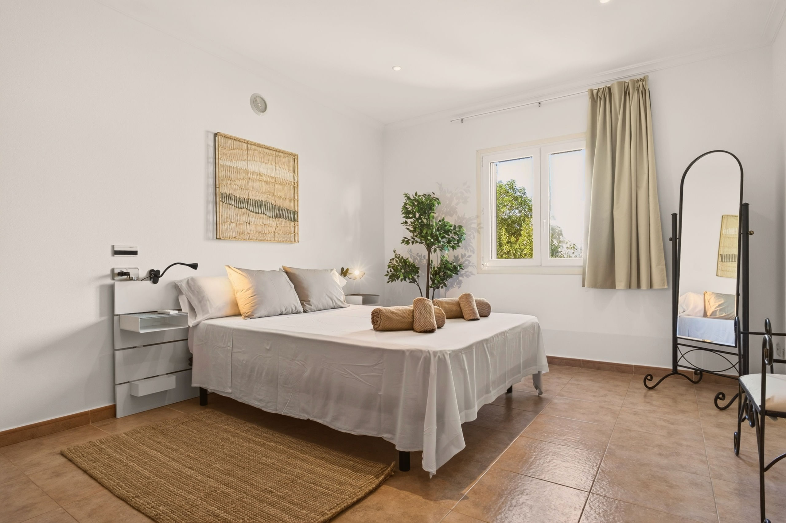 Zweites Doppelschlafzimmer mit grauer Bettwäsche, rechteckigem Wandteppich aus Naturfasern, Fenster mit beigem Vorhang, Jute-Teppich und hohem Standspiegel.