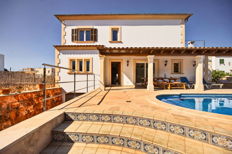 Weiß verputzte zweistöckige Villa mit hellen Naturstein-Ecken, davor Naturstein-Terrasse, Fliesenstufen mit bemalten Kacheln, rechts Poolrand und überdachte Terrasse mit Tisch und Liegen.
