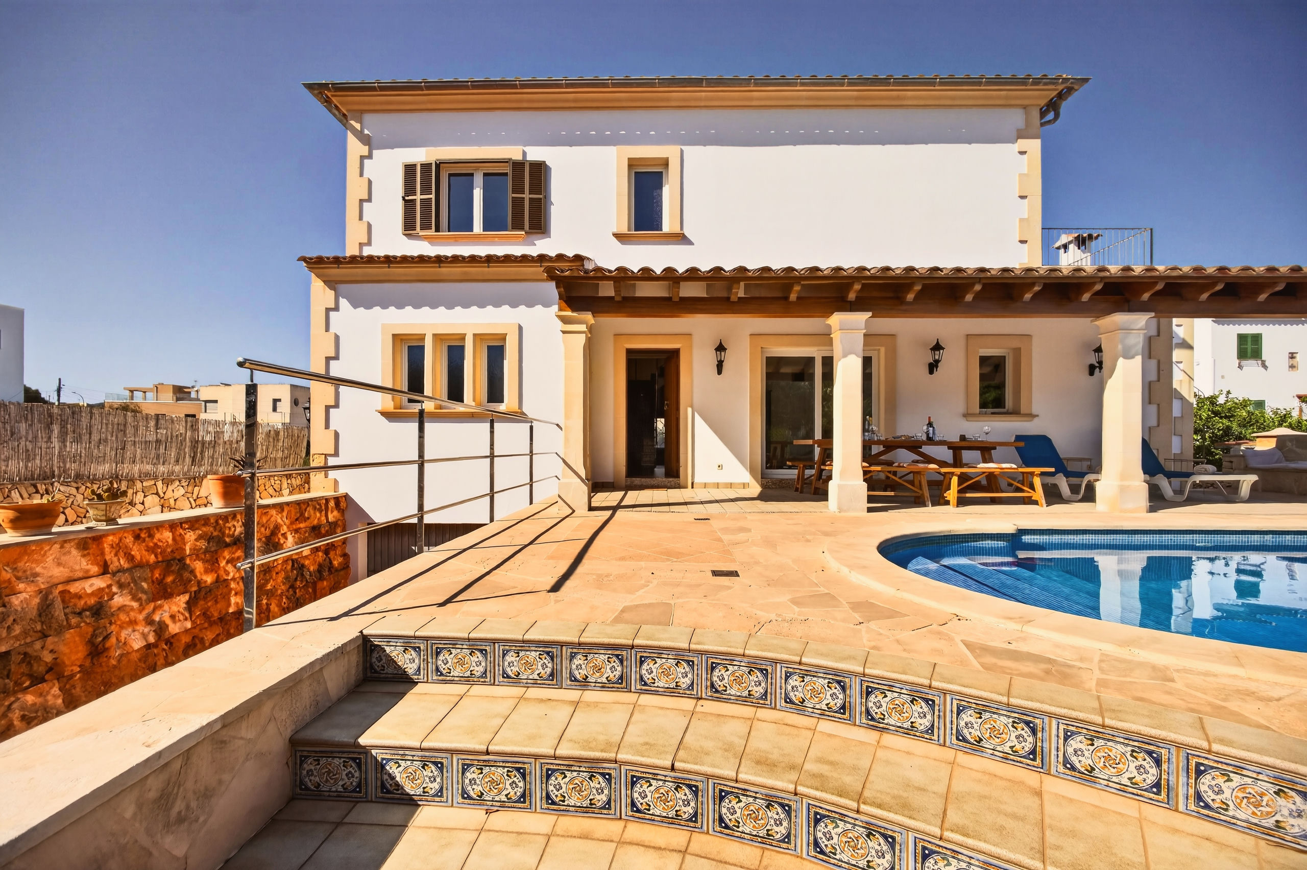 Weiß verputzte zweistöckige Villa mit hellen Naturstein-Ecken, davor Naturstein-Terrasse, Fliesenstufen mit bemalten Kacheln, rechts Poolrand und überdachte Terrasse mit Tisch und Liegen.