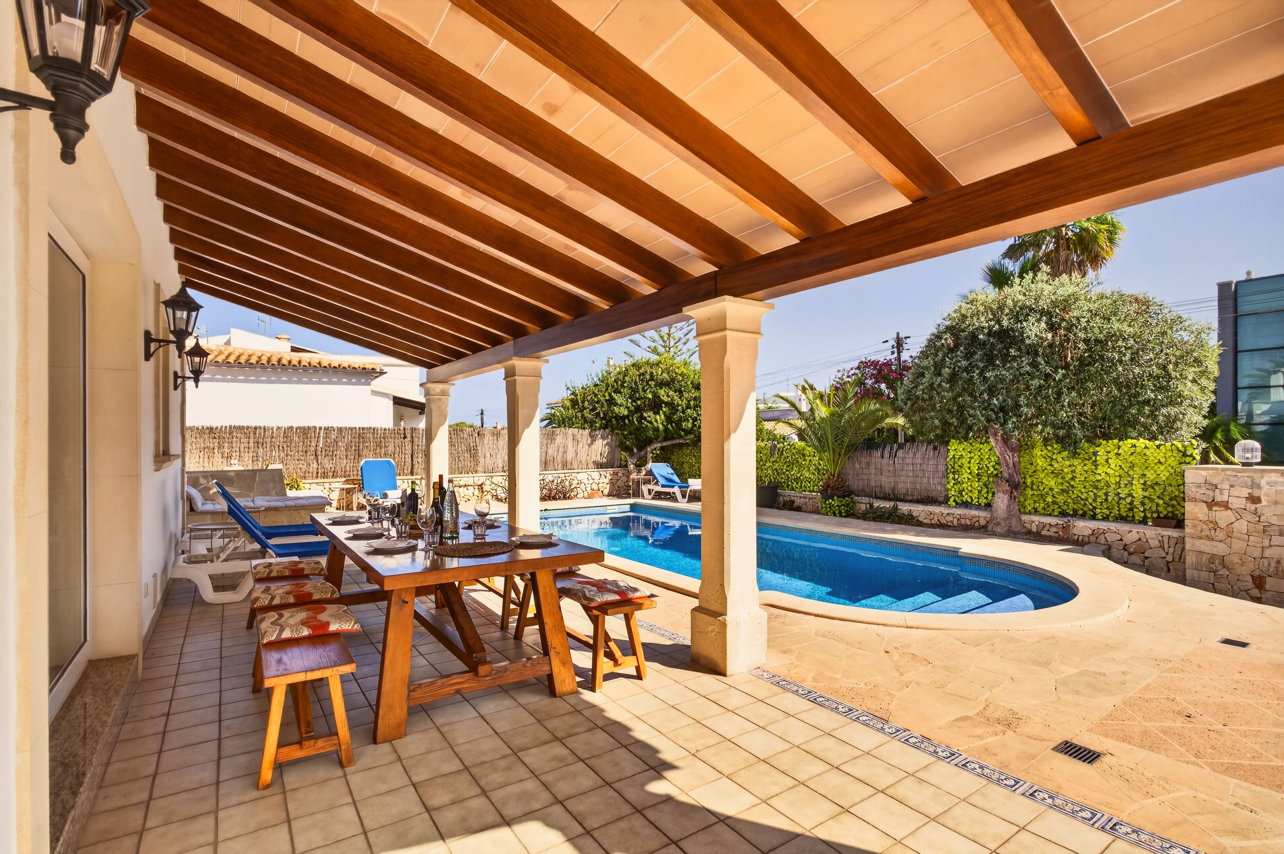 Unter der Holzpergola steht eine lange Holztafel mit Hockern gedekt bereit, dahinter der Pool zwischen Hecken, Olivenbaum und Palmen.
