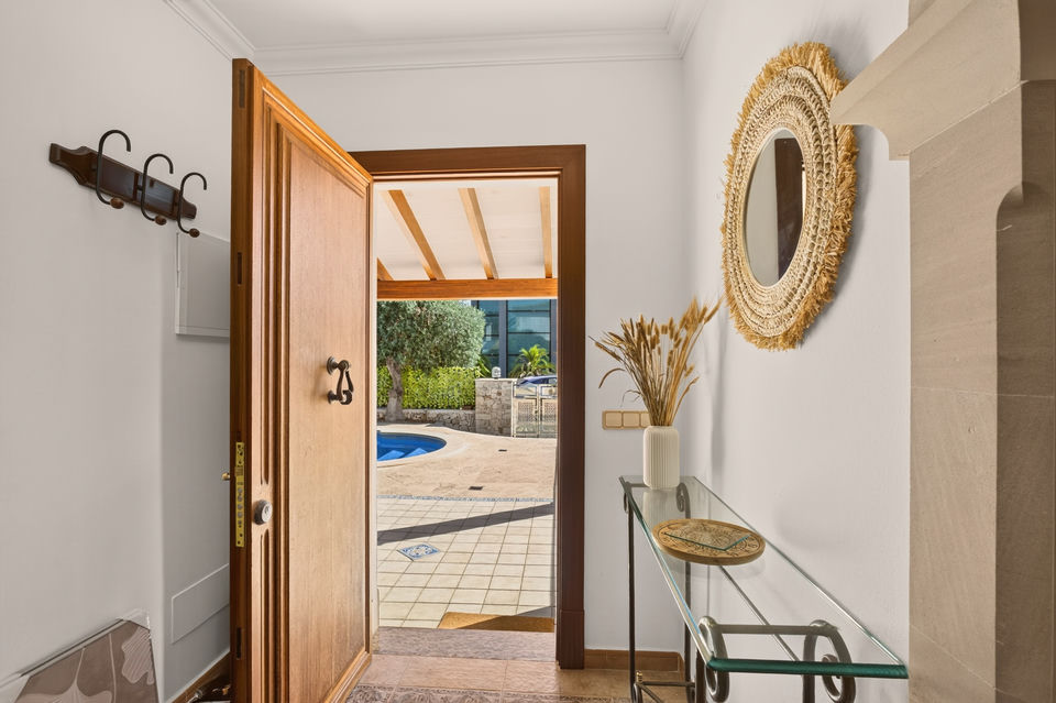 Diele mit offener Holztür, dahinter der geflieste Patio mit Poolrand; runder Rattanspiegel an der Wand, Glas-Konsolentisch mit Trockenblumen, Garderobenhaken links.