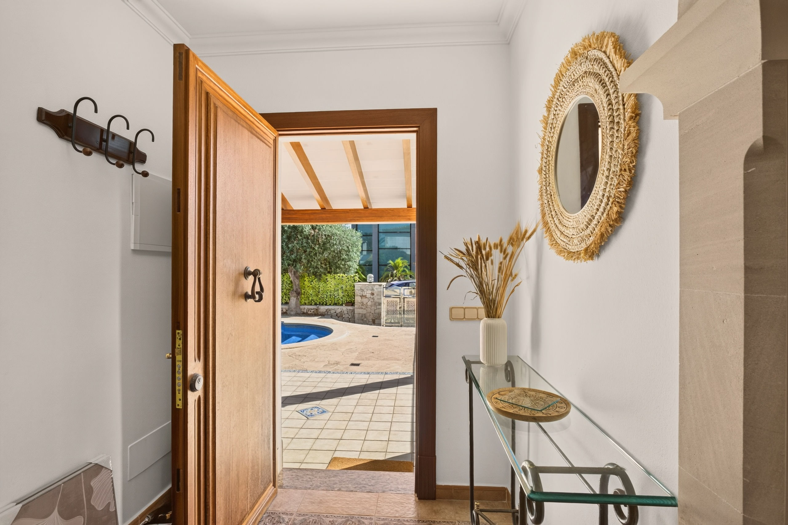 Diele mit offener Holztür, dahinter der geflieste Patio mit Poolrand; runder Rattanspiegel an der Wand, Glas-Konsolentisch mit Trockenblumen, Garderobenhaken links.
