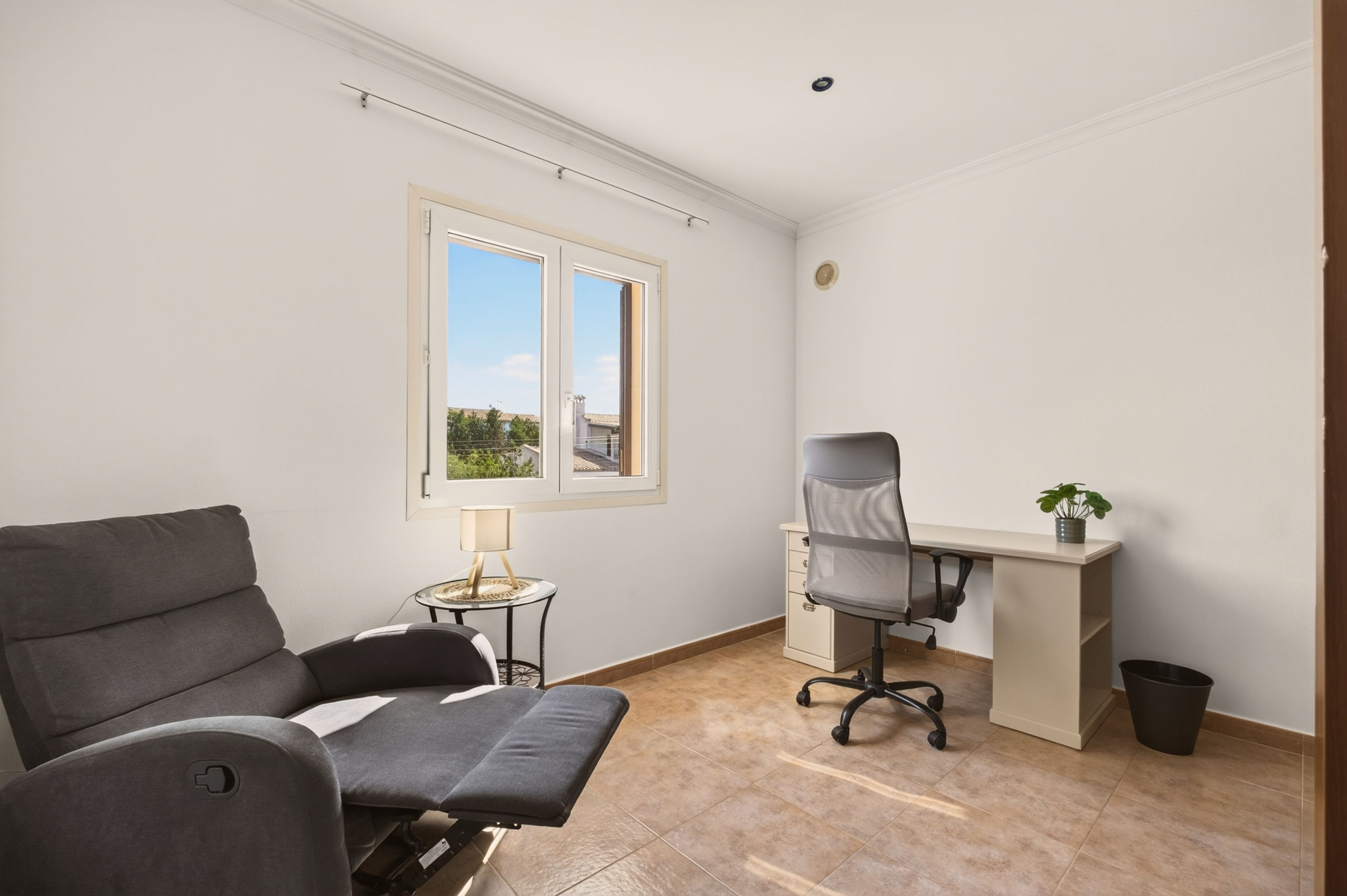 Kleines Arbeitszimmer mit grauem Relaxsessel, Glastisch mit Lampe, Schreibtisch mit Netz-Drehstuhl, kleiner Pflanze und Fenster zum Blauen.