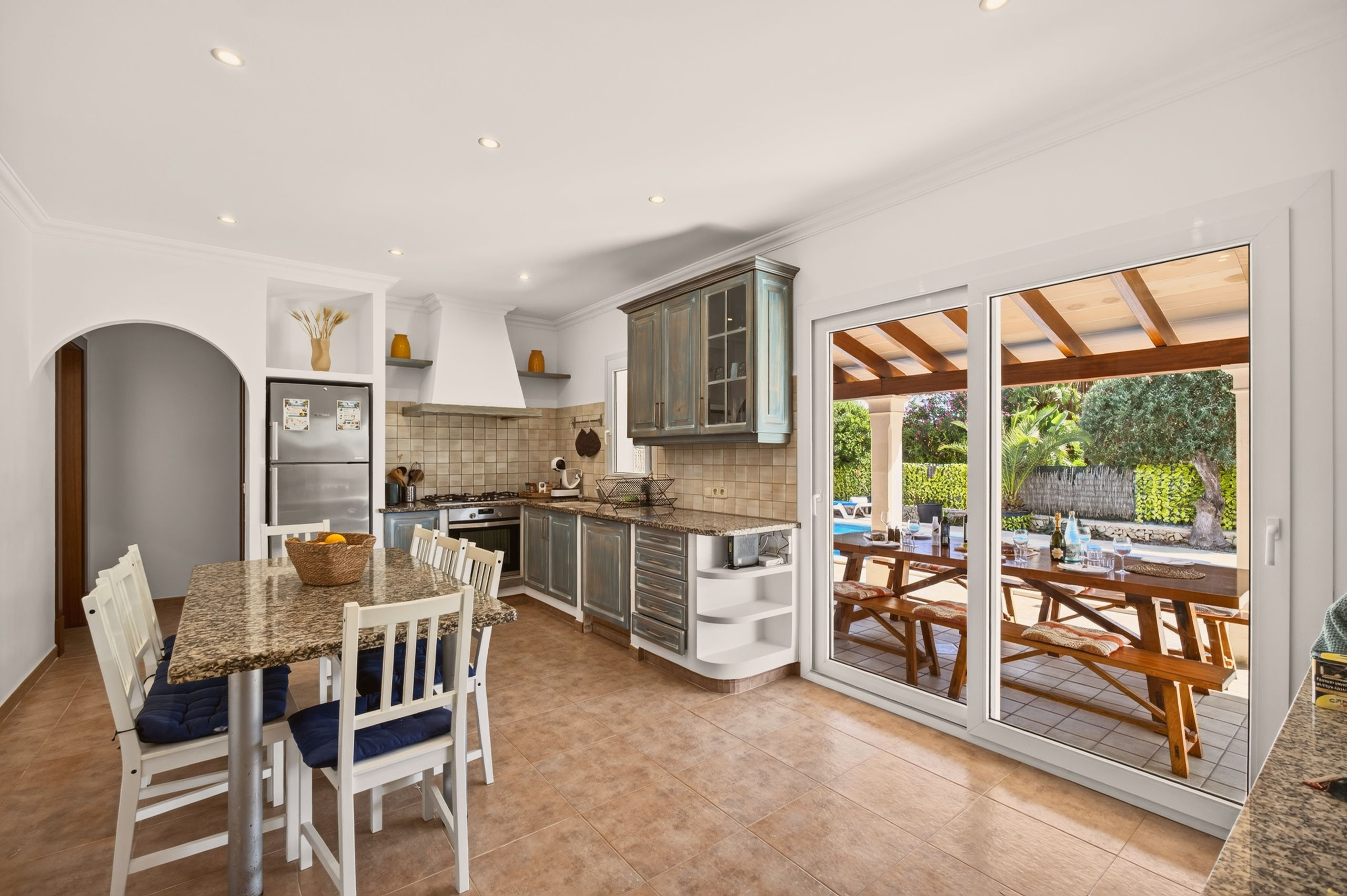 Wohnküche mit Granittisch und weißen Stühlen, blau-grauen Landhaus-Schränken, Fliesenspiegel und Schiebetüren zur überdachten Terrasse.