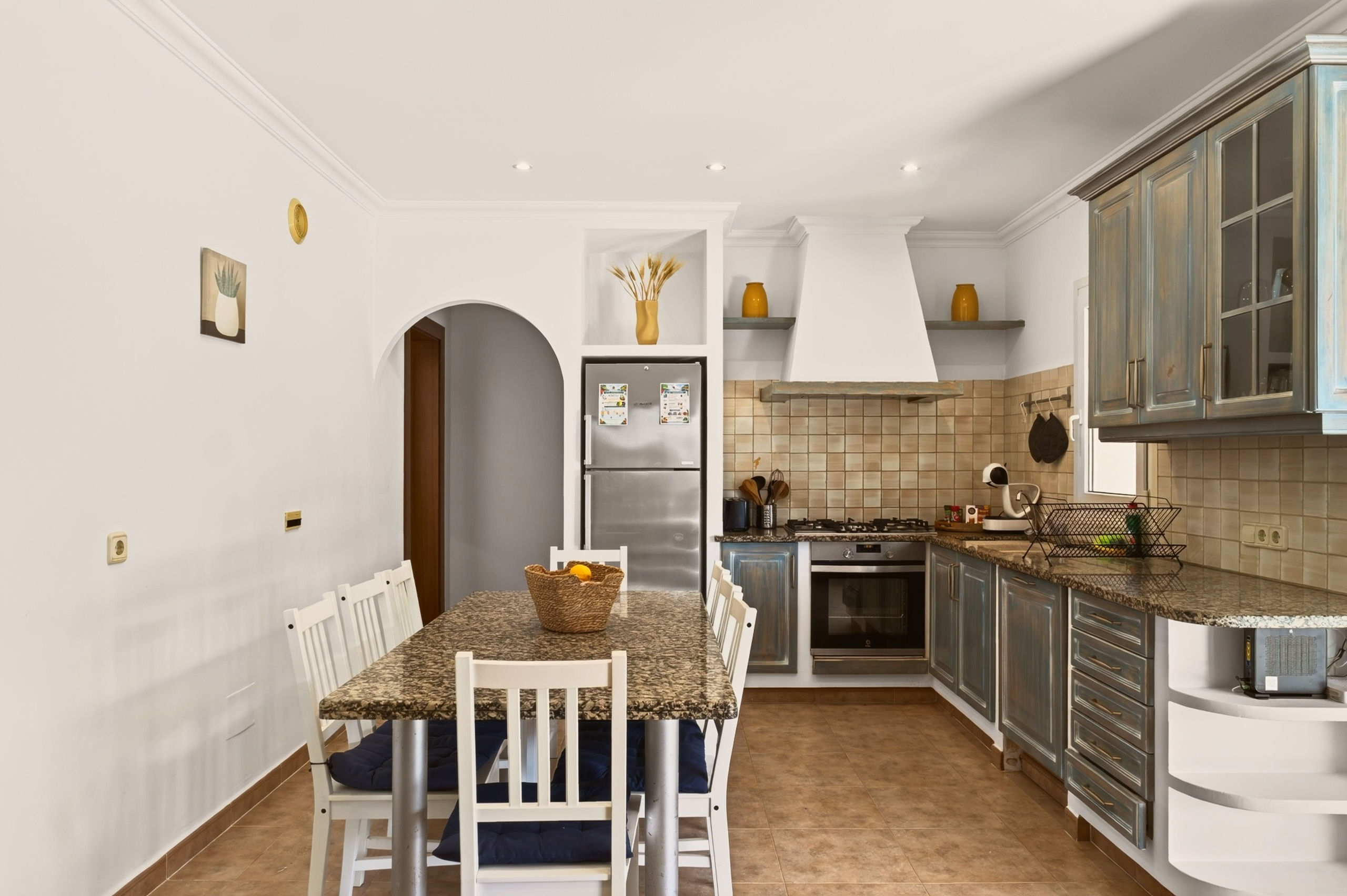 Blickfang in die Küche: Granittisch mit Obstkorb, bogenförmiger Durchgang, Nische mit Kühlschrank, Ecke mit Herd und weißer Haube, blaue Holzfronten.