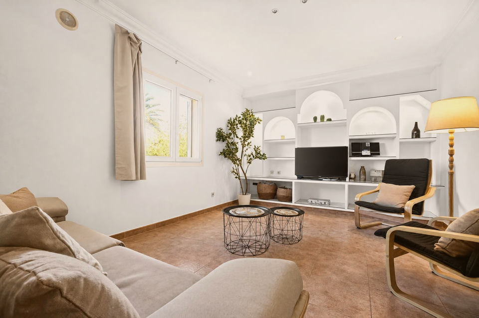 Wohnzimmer mit beigen Sofas, schwarzen Draht-Beistelltischen, zwei Holzsesseln mit dunklen Polstern und Fernseher in weißer Regalwand mit Nischenbögen.