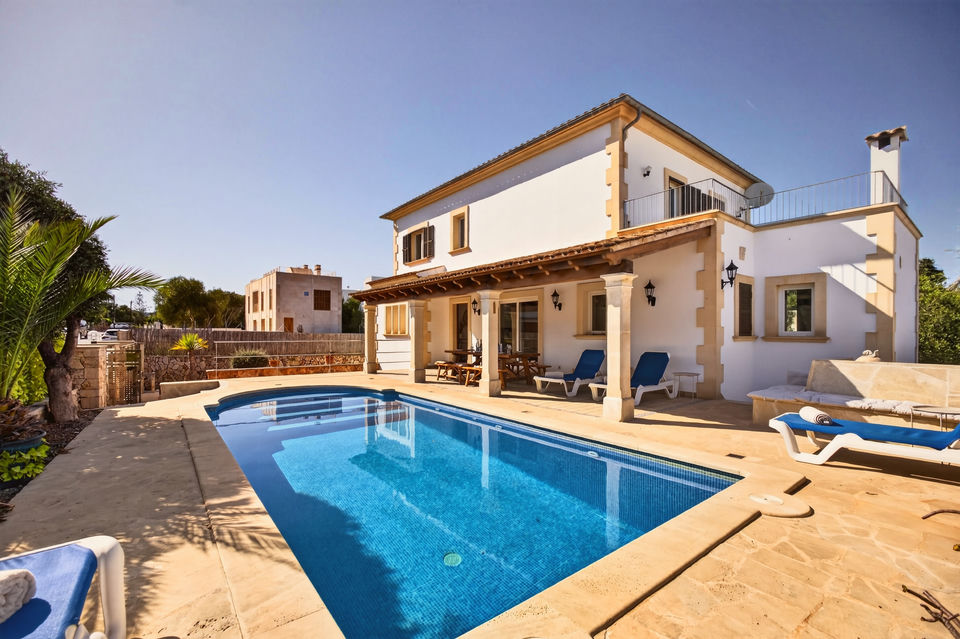 Erhöhte Ansicht des frei geformten türkisfarbenen Pools mit Natursteinterrasse, Sonnenliegen, Sonnenschirm und Essgruppe, dahinter das weiß getünchte Haus mit Turm zwischen Palmen.